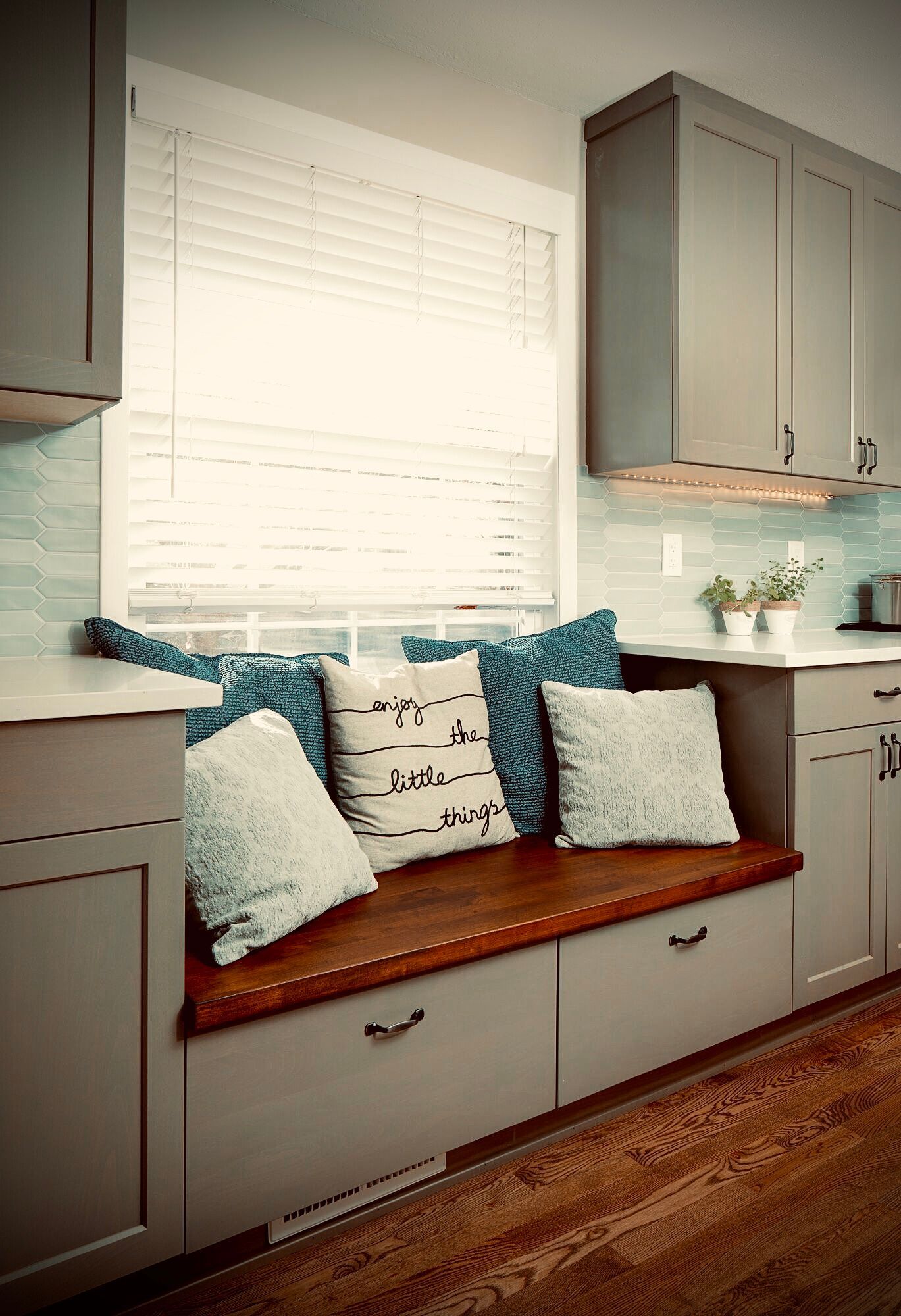 Kitchen with window seat, gray cabinets, wooden bench, blue and white pillows, blinds, white countertop.