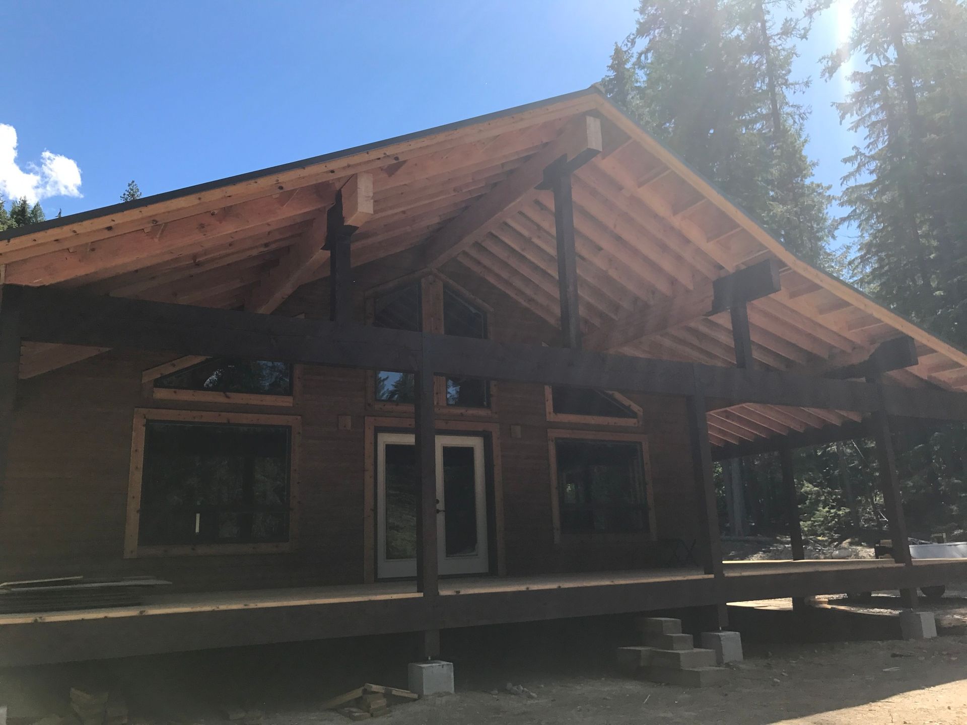Wooden cabin under construction with a porch and exposed beams; set in a forest on a sunny day.