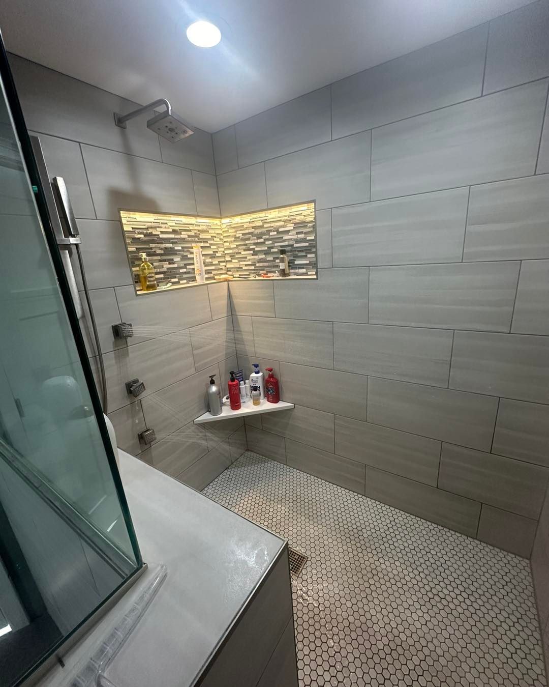 Modern bathroom shower with gray tile walls and pebble floor.  Shelf holds bottles. Glass shower door on left.