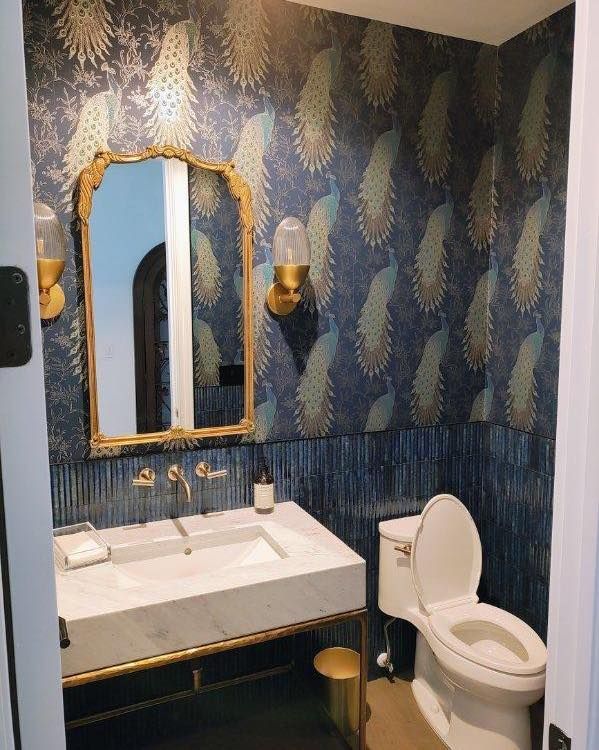 A small bathroom with patterned blue and gold wallpaper, gold mirror, sink, toilet, and sconces.