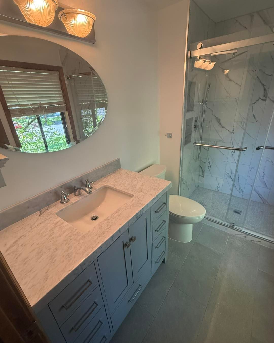Bathroom with marble-look vanity, round mirror, glass shower, and white toilet. Light blue cabinet and gray floor.