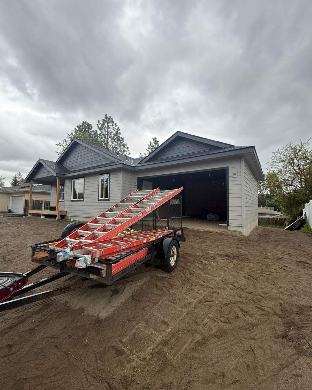 New construction house with open garage. A red ladder on a trailer parked on a dirt lot. Overcast sky.