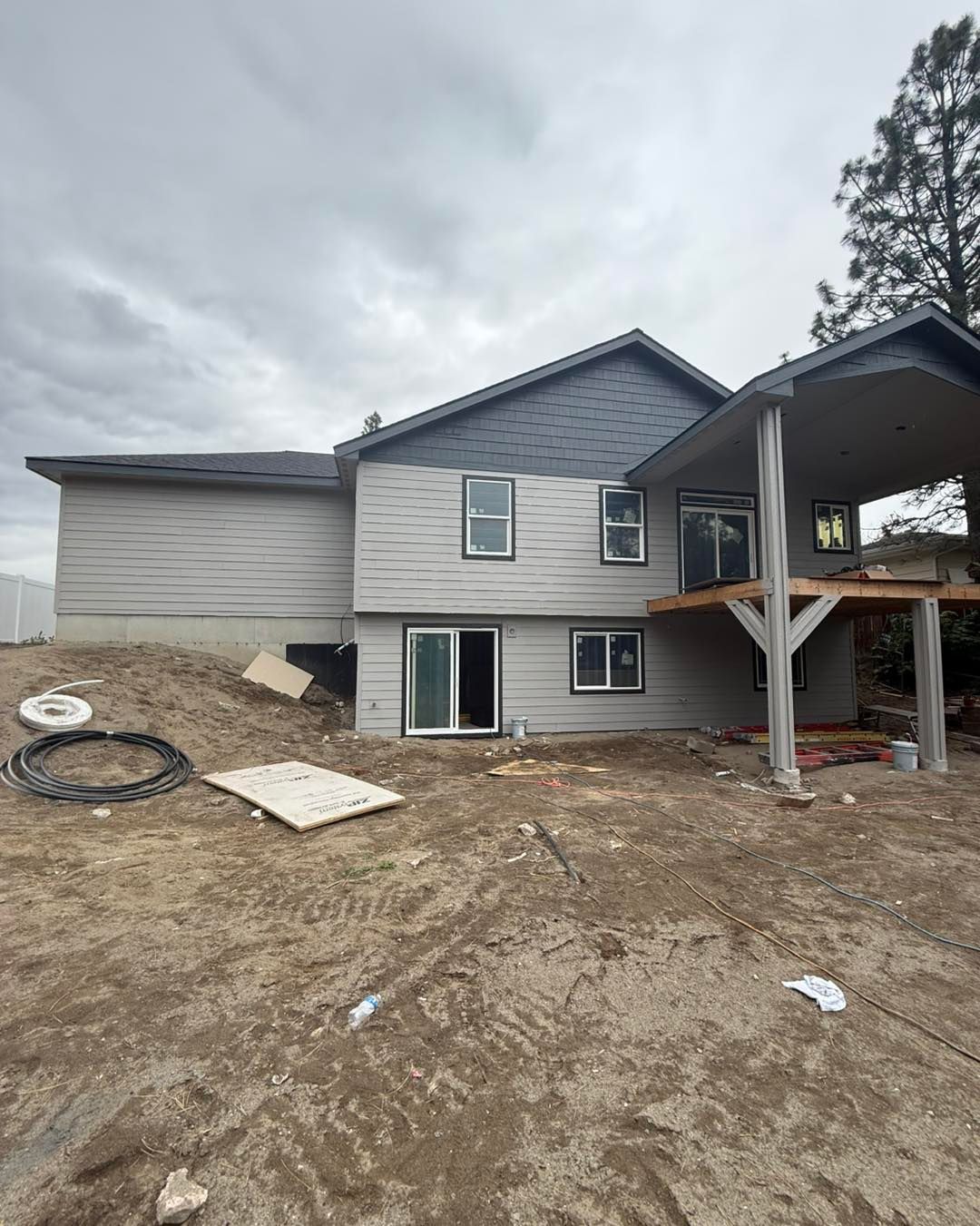 Exterior of a house under construction. Gray siding, roof, and a covered patio on a dirt lot under an overcast sky.