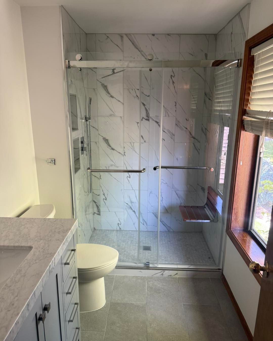 Bathroom with marble-like tile shower, sliding glass doors, a toilet, and a window with blinds.