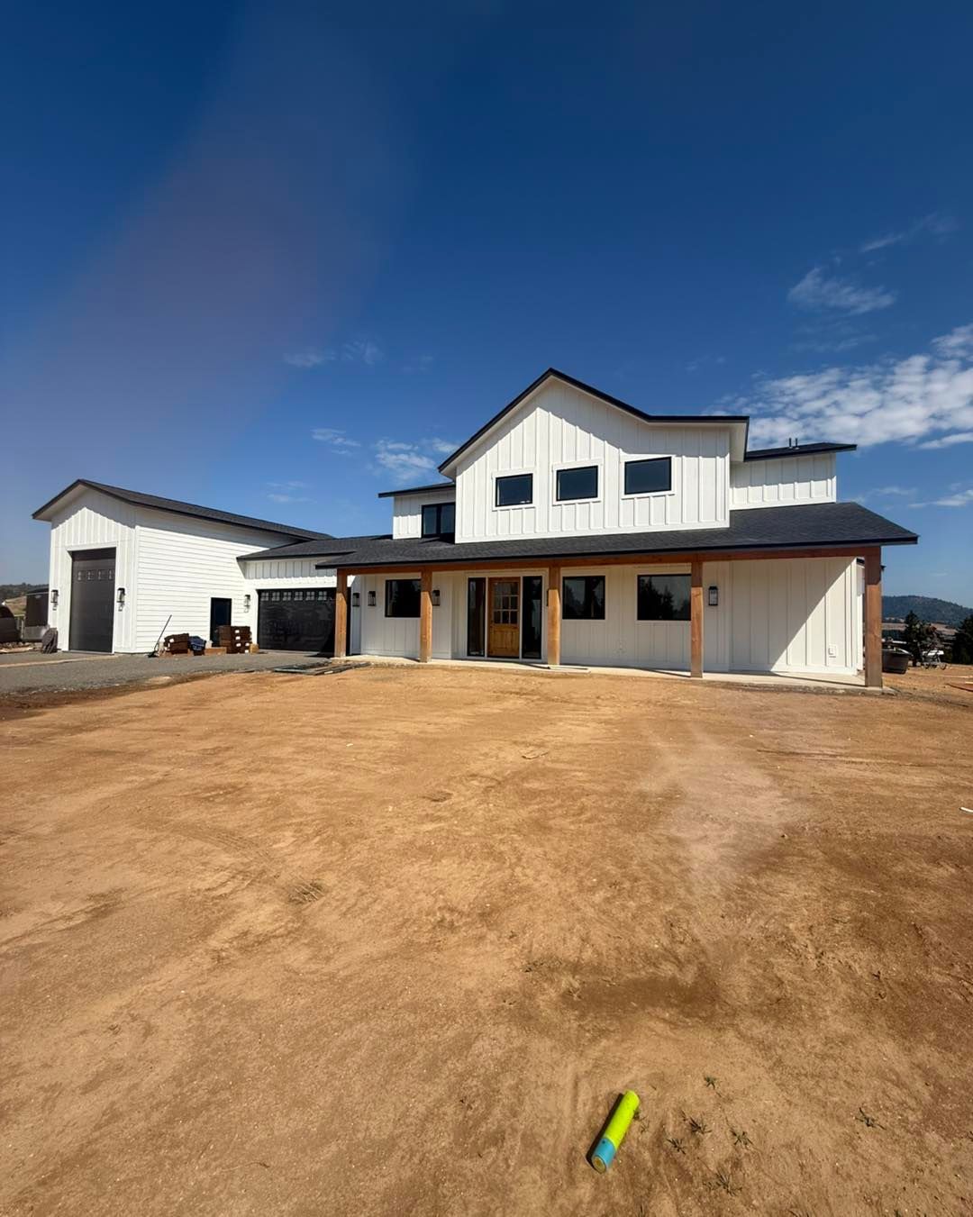 White farmhouse with black trim under a blue sky, on a dirt lot. Garage on the left.
