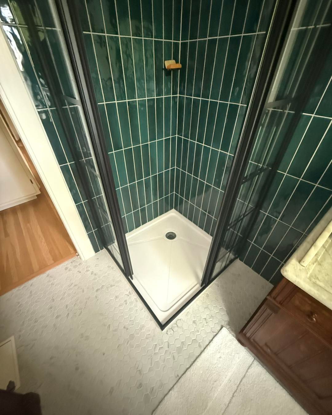 Corner shower with green tiled walls and a white base, next to a wood cabinet and open door.