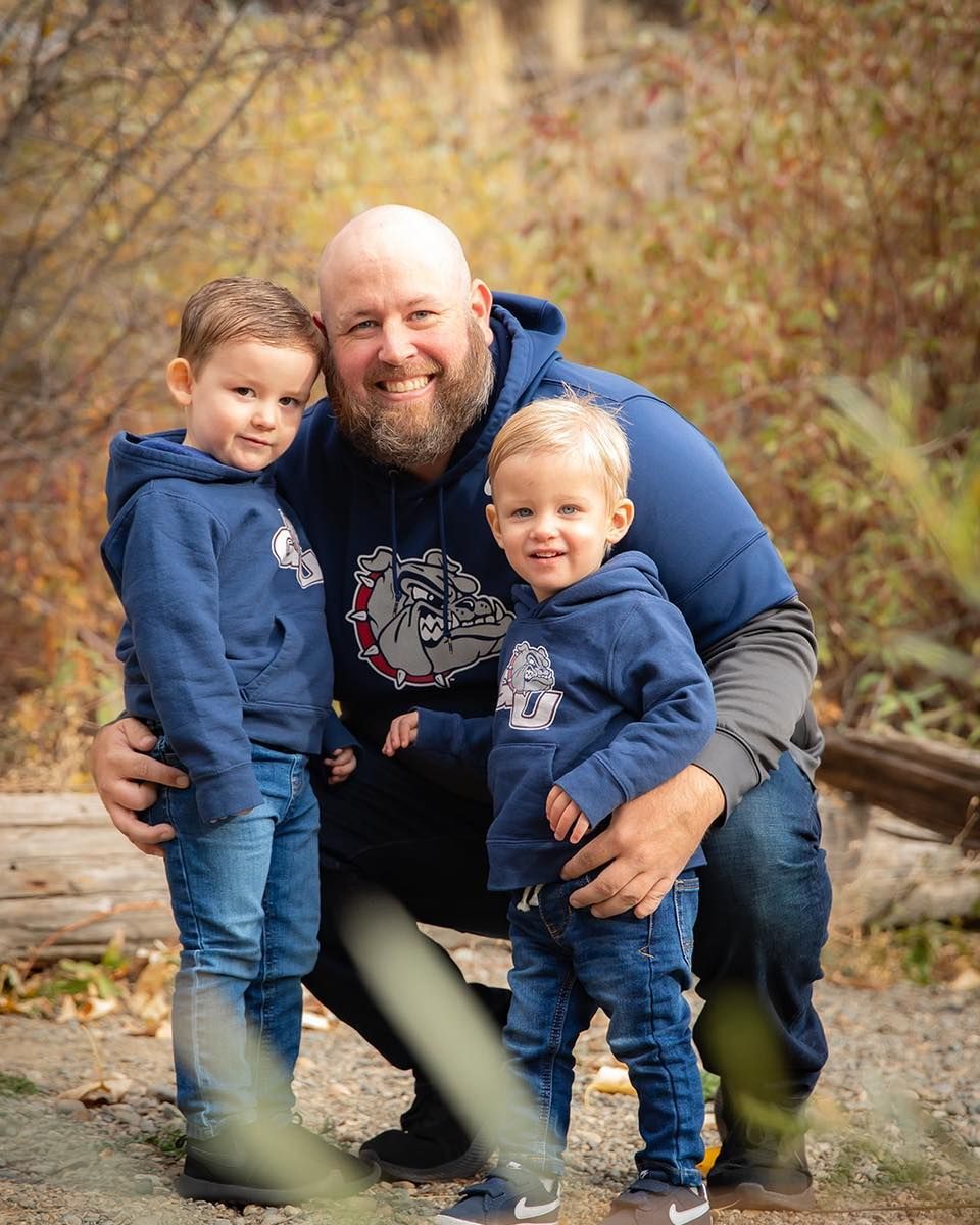 Man kneels with two young children. All wear blue hoodies and jeans outdoors. Autumn foliage.