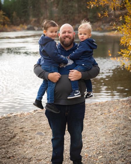 Man holding two young children near a lake; all wearing blue hoodies and jeans. Fall foliage in background.