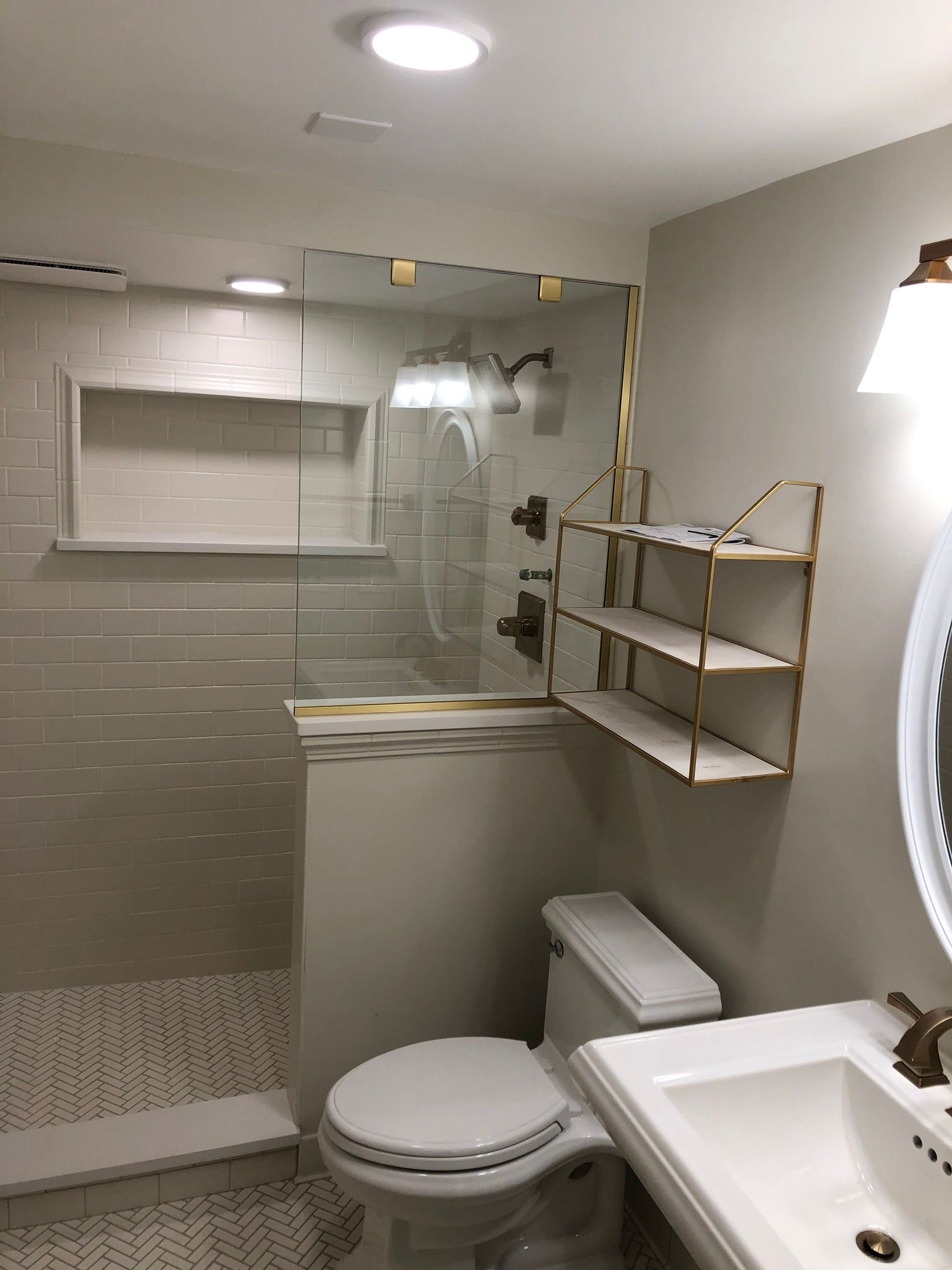 Bathroom with glass shower, toilet, sink, shelving, and off-white and gold accents.