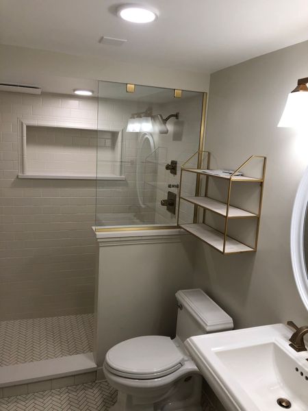 Bathroom with glass shower, toilet, sink, shelving, and off-white and gold accents.