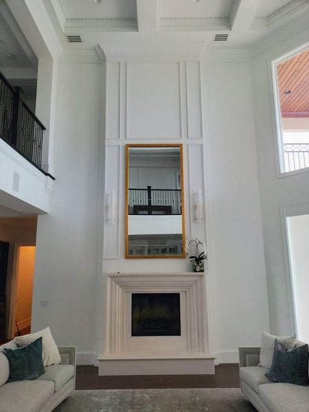 Tall white fireplace with gold-framed mirror, flanked by two couches, high ceiling with molding.