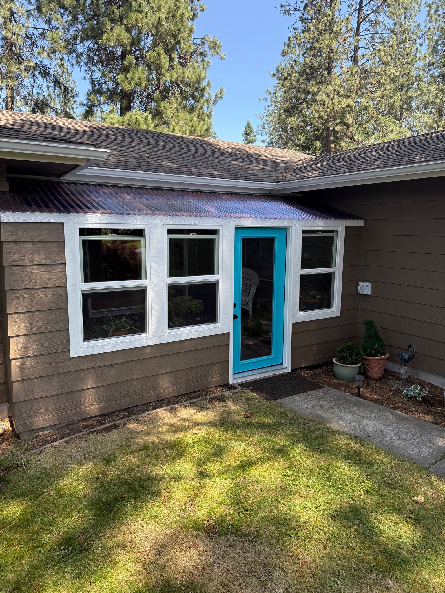 Brown house with white-trimmed windows and teal door under a corrugated awning. Green grass in foreground.