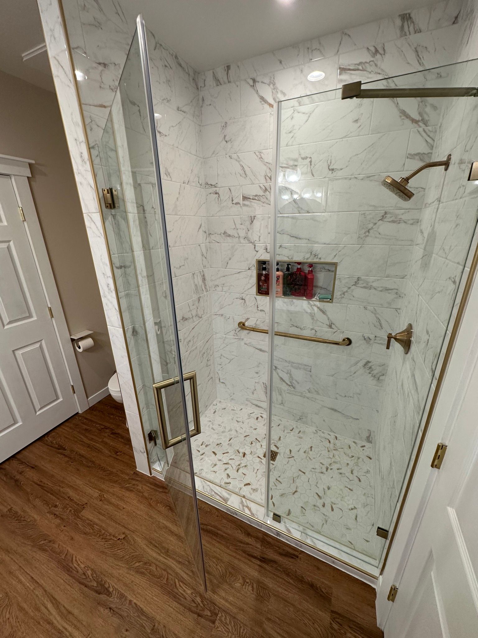 Bathroom with marble tile shower, glass door, and gold fixtures. Wooden floor.
