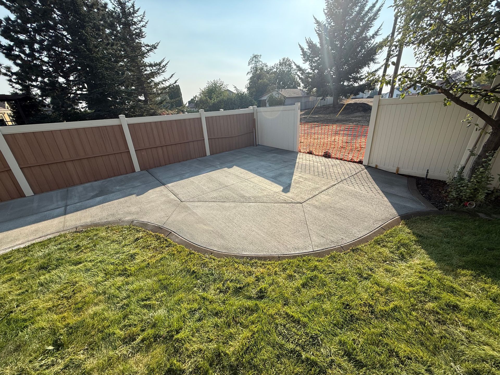 Lawn and concrete patio surrounded by fences, trees, and bright sunlight.