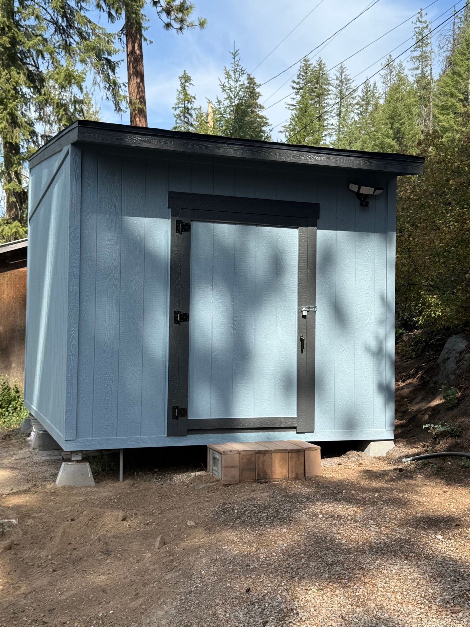 Blue shed with black trim and a security camera, set on concrete blocks outdoors.