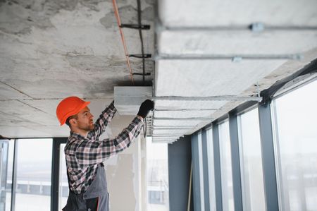 Construction worker in hard hat installs ceiling panel, working in a building with large windows.