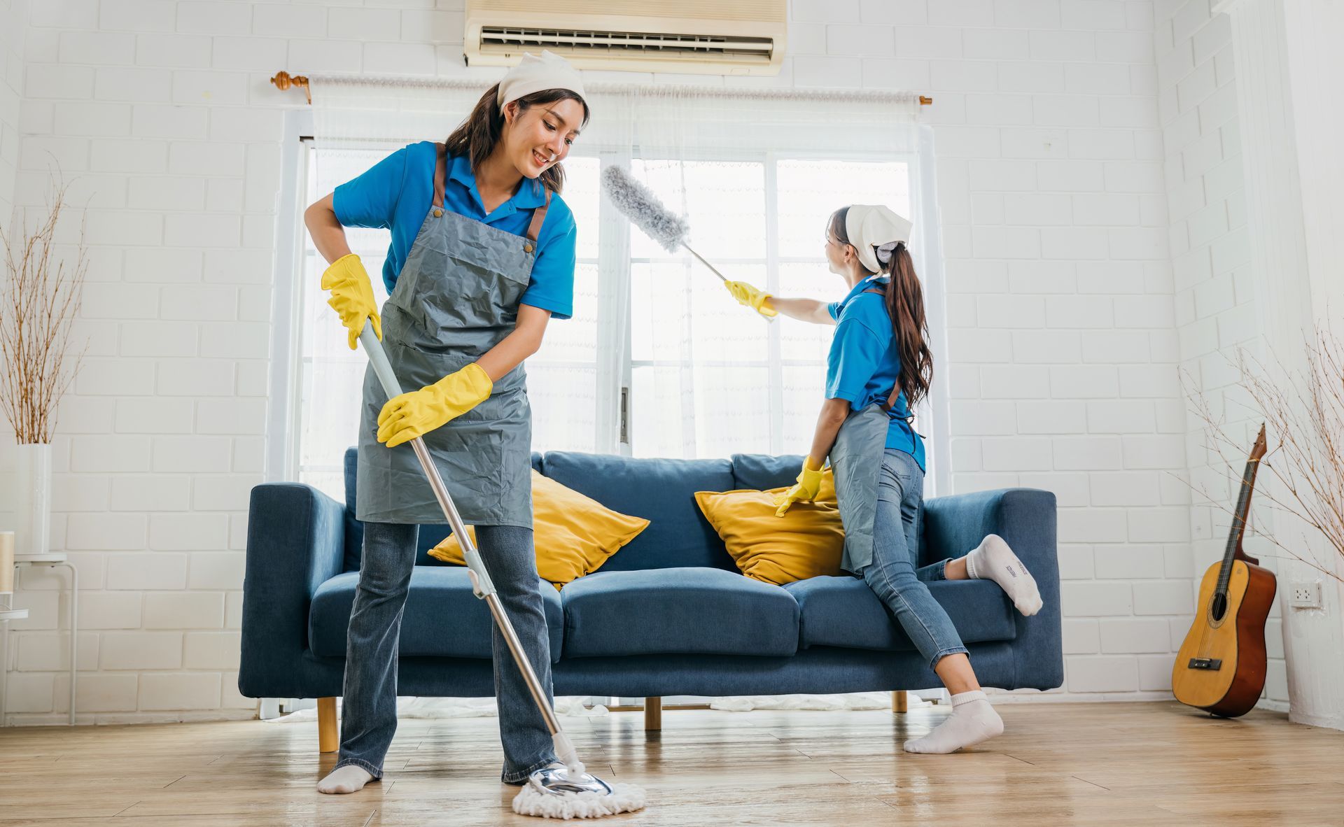 Two people in blue shirts, aprons, and head coverings clean a room; one mops the floor while the other dusts the window.