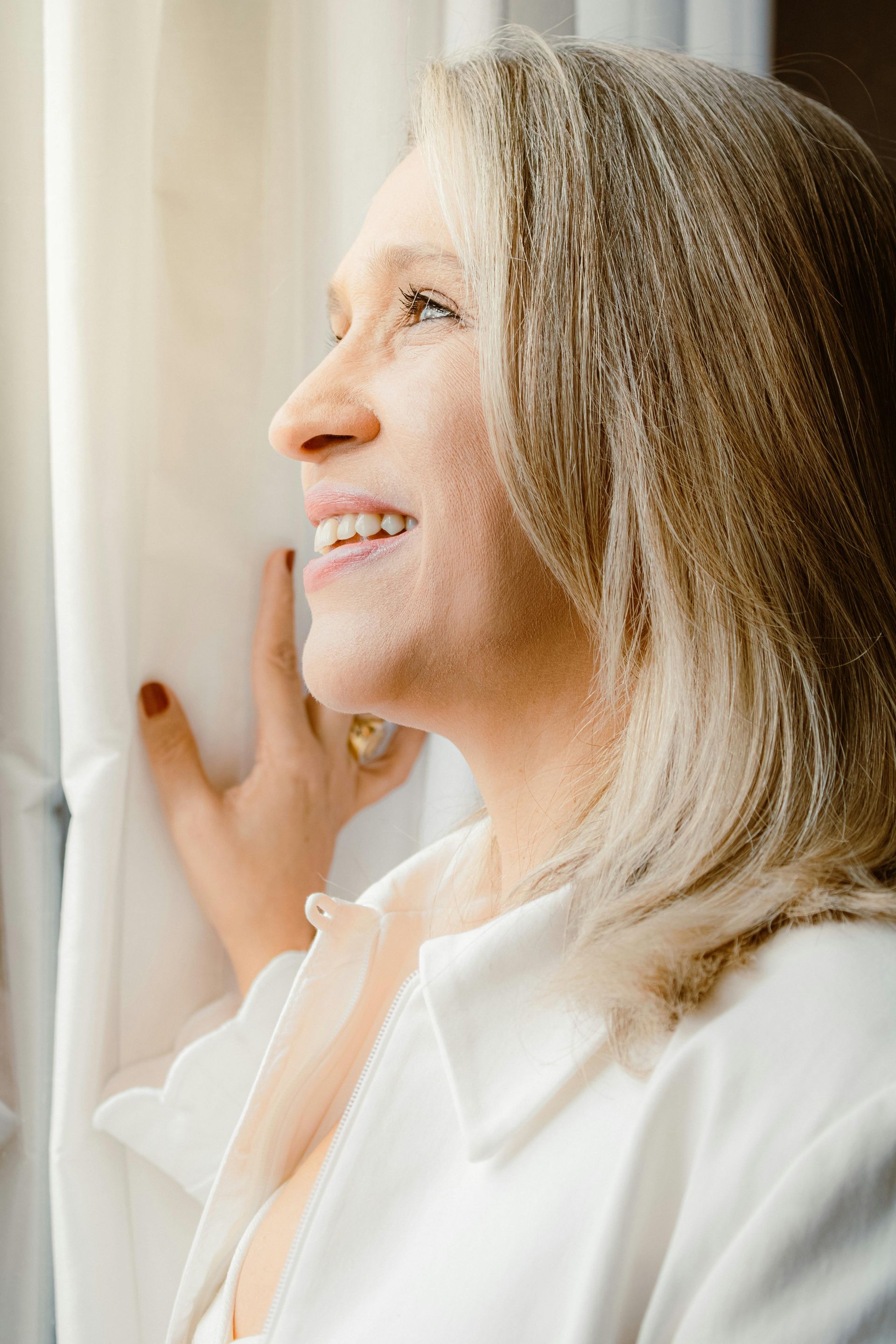 A woman in a white shirt is smiling while looking out of a window.