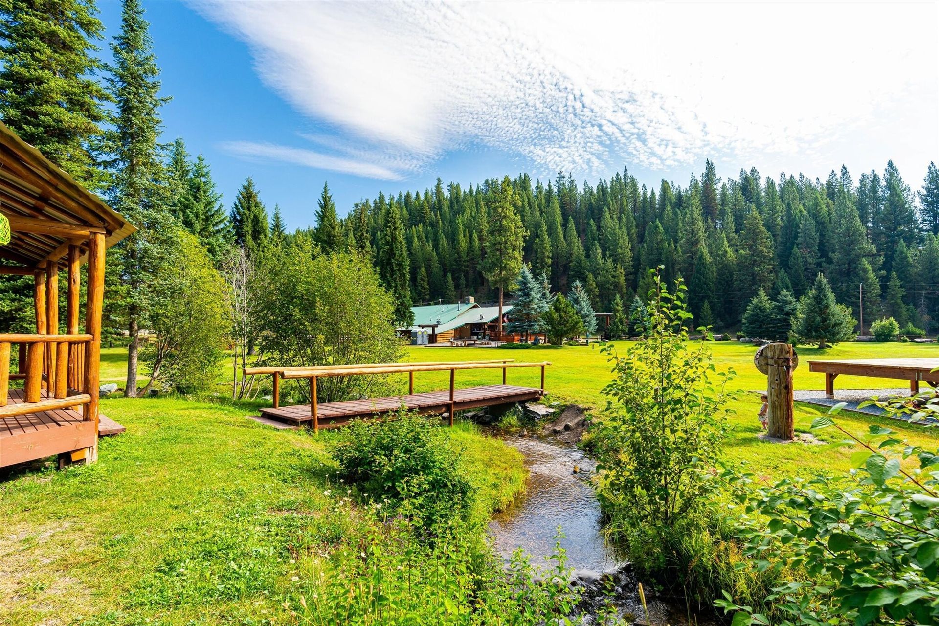 Wooden cabin beside a stream and bridge in a lush green forest meadow under a blue sky