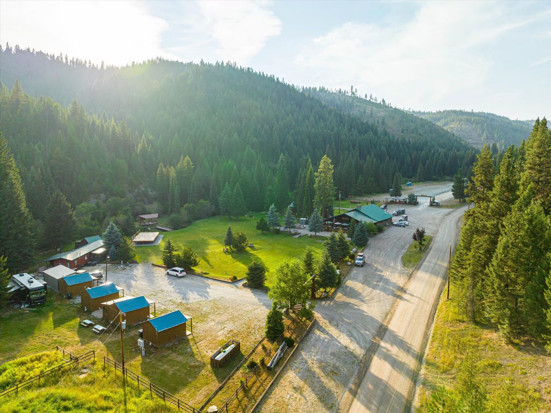 Aerial view of a sunny forest campground with cabins, tents, and a winding dirt road beside hills.