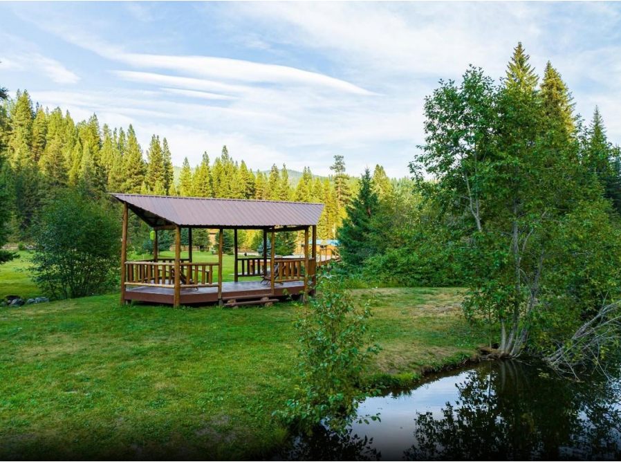 Wooden gazebo in a lush green meadow beside a small pond, surrounded by forest and hills.