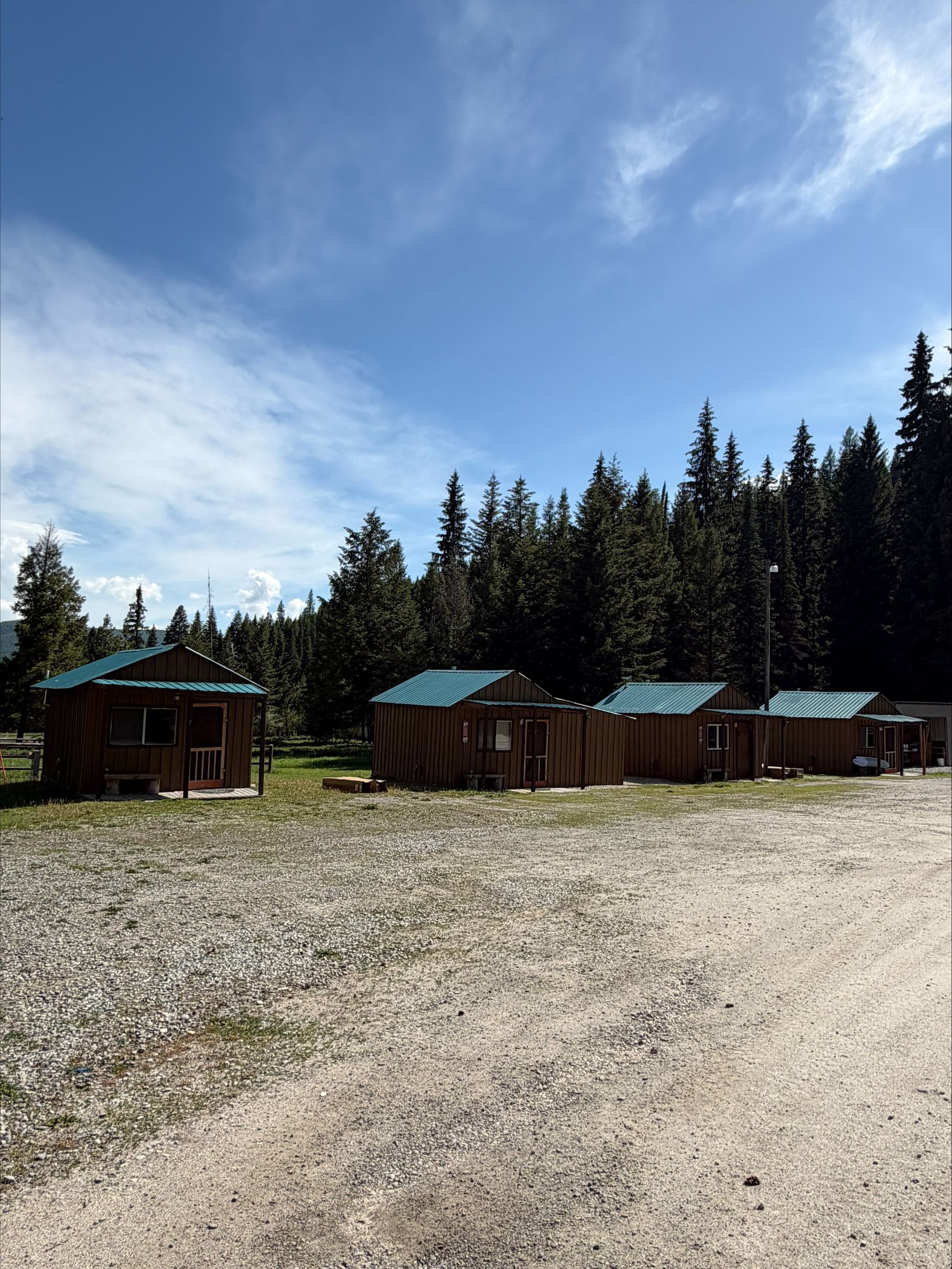 Rustic cabins with green roofs beside a gravel clearing and pine forest under a blue sky