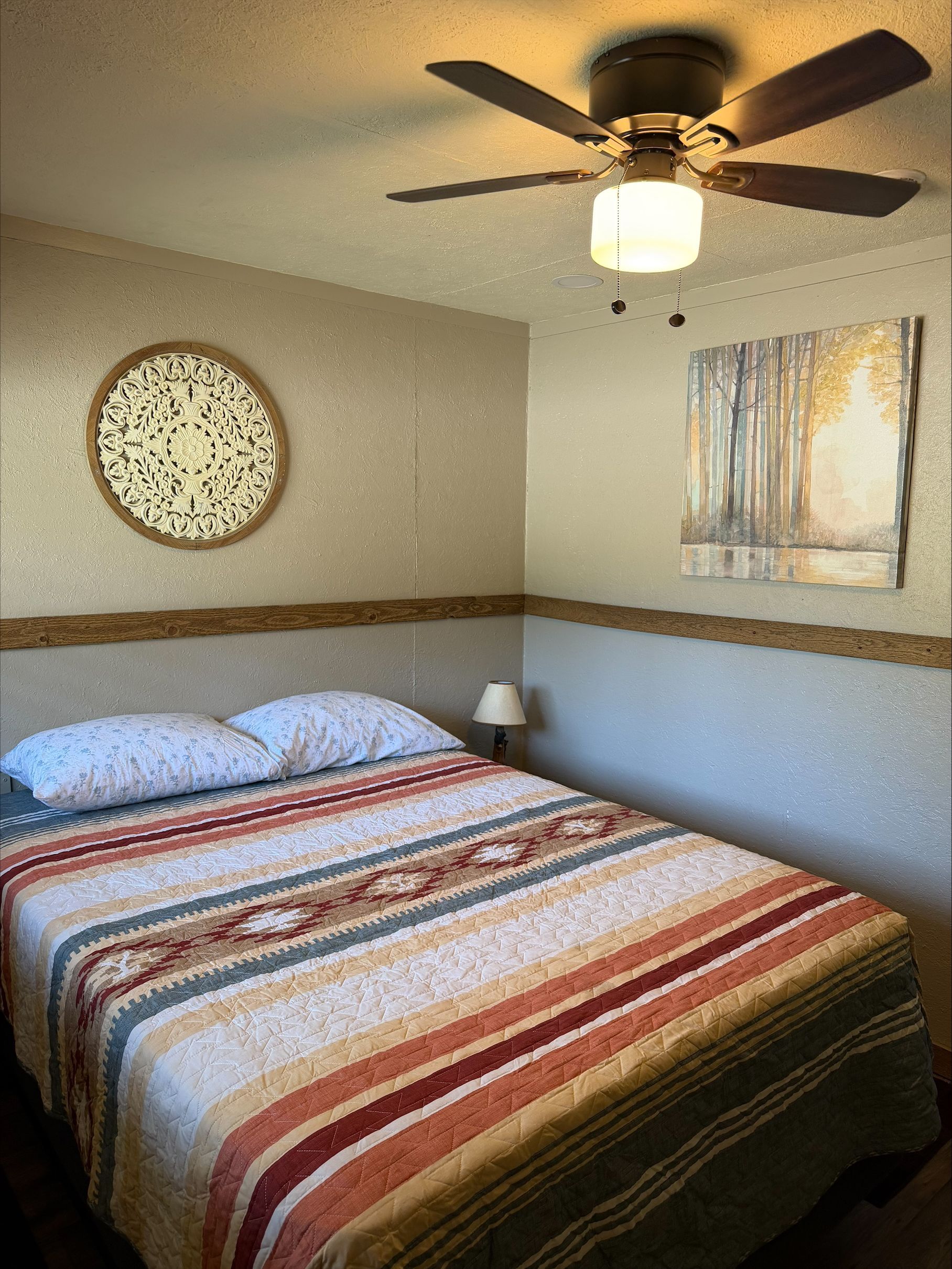 Cozy bedroom with striped bedspread, ceiling fan, round wall decor, and a small window with curtains.