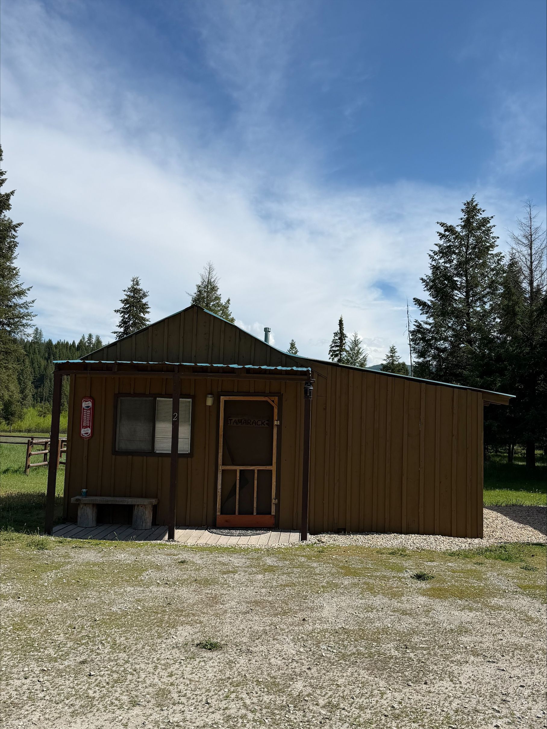 Small rustic brown cabin with a tin roof in a gravel clearing under a blue sky, surrounded by trees