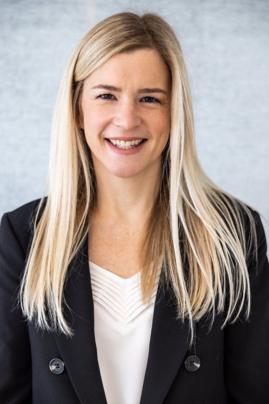 A woman with long blonde hair is wearing a black jacket and smiling.