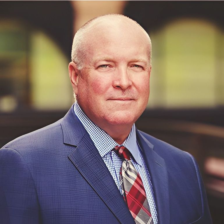 Portrait of a bald man in a blue suit and red plaid tie against a softly blurred background