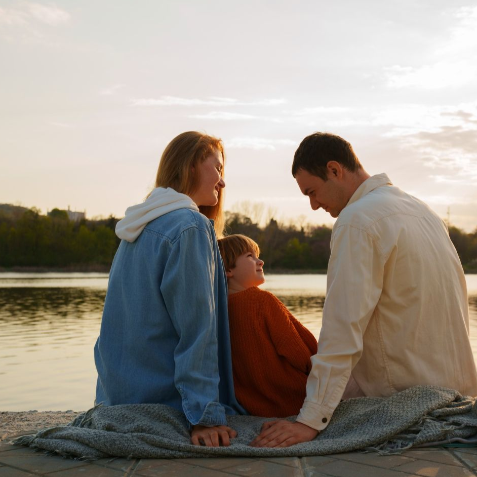 Three people sit on a blanket by a lake at sunset, facing each other and smiling.