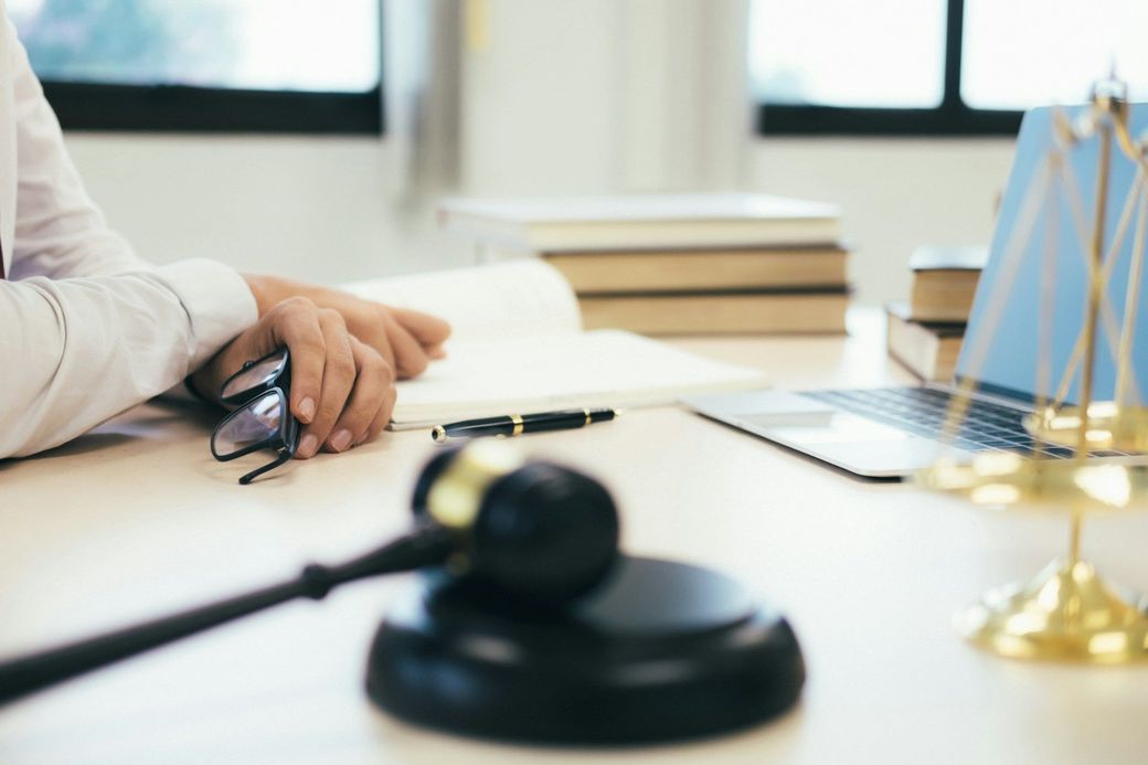 Lawyer’s desk with gavel, folded hands, laptop, and scales of justice in a bright office