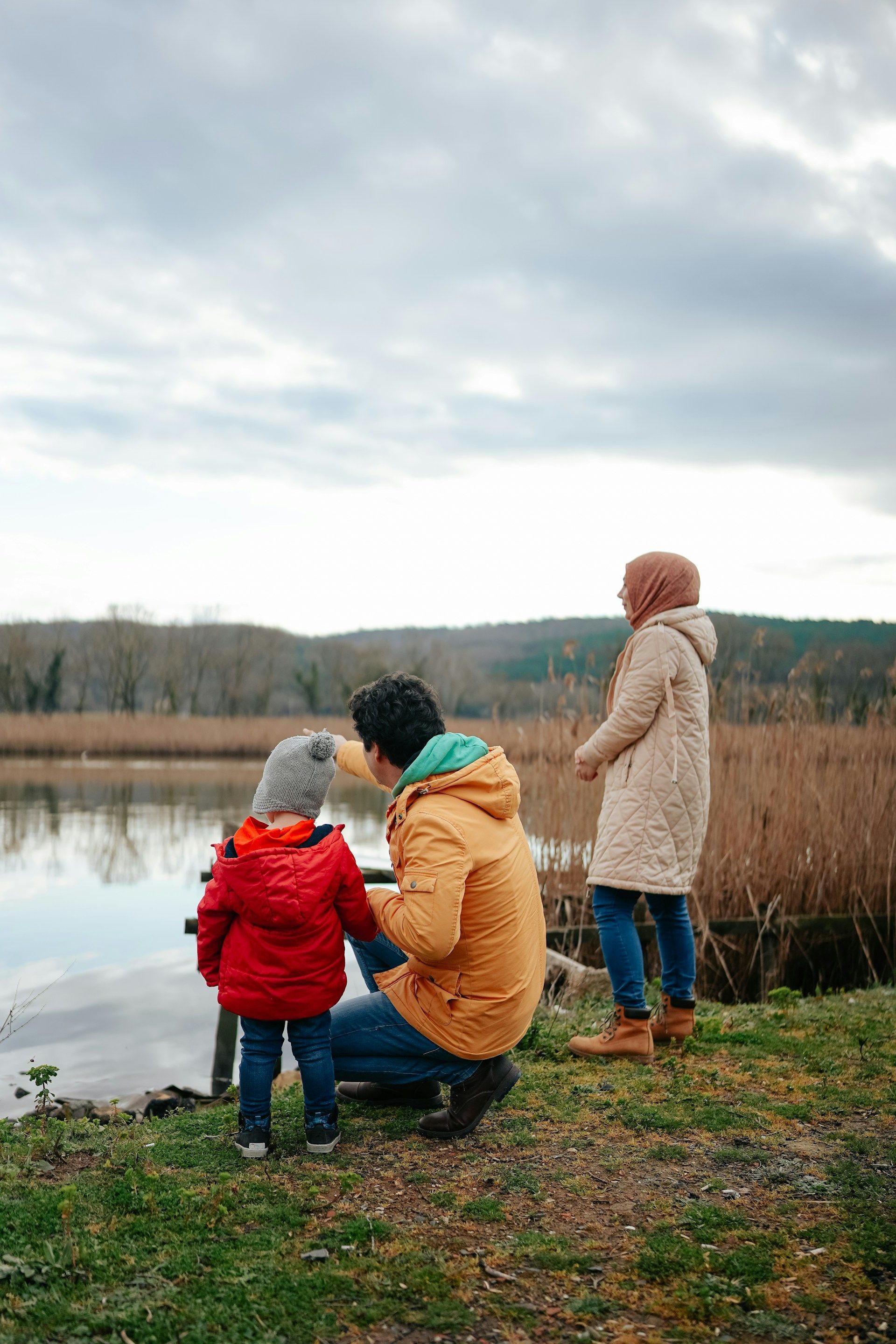 Three people in winter coats by a lakeside, looking out over calm water under a cloudy sky