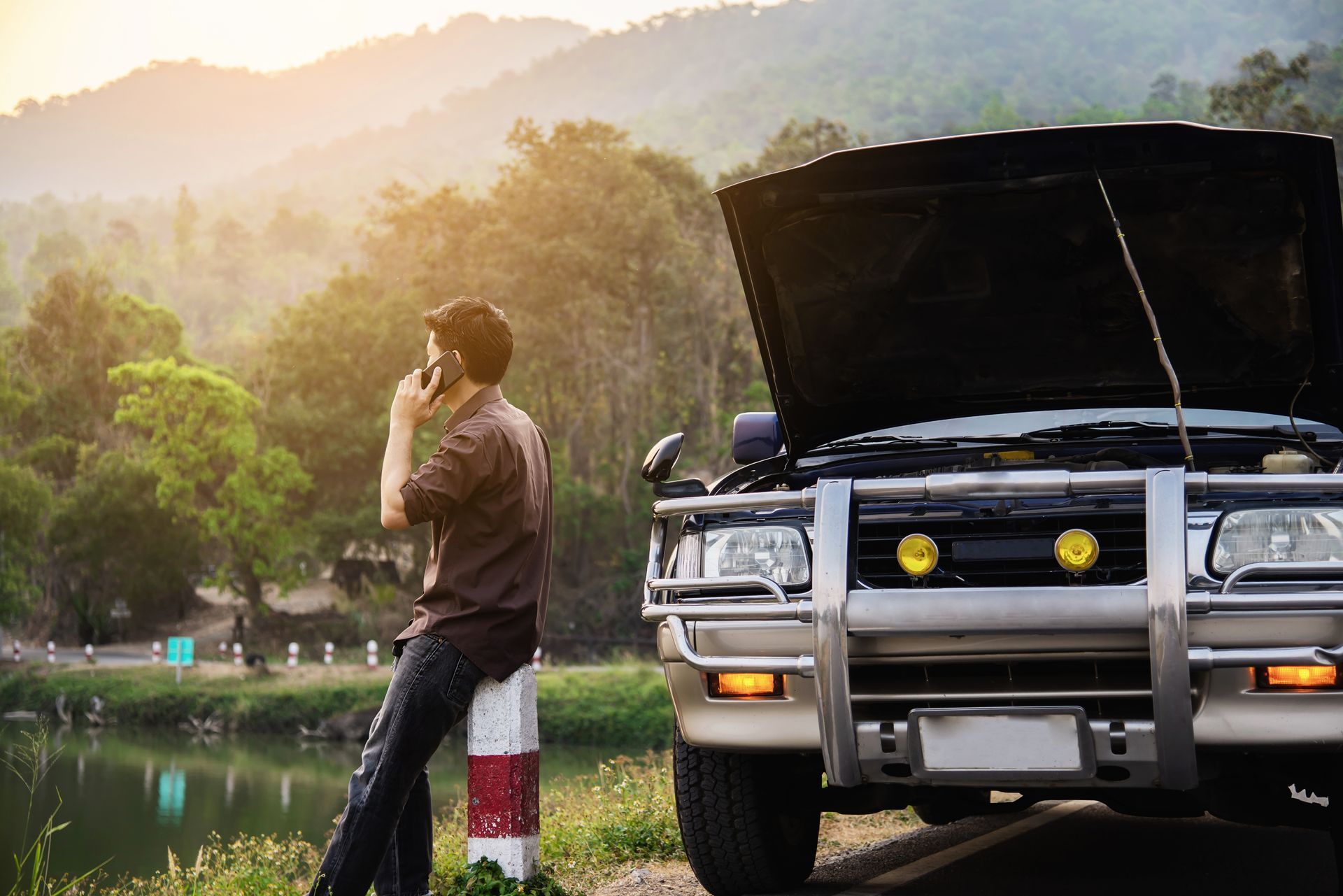 Person leaning on roadside marker beside open-hood SUV near a lake at sunset
