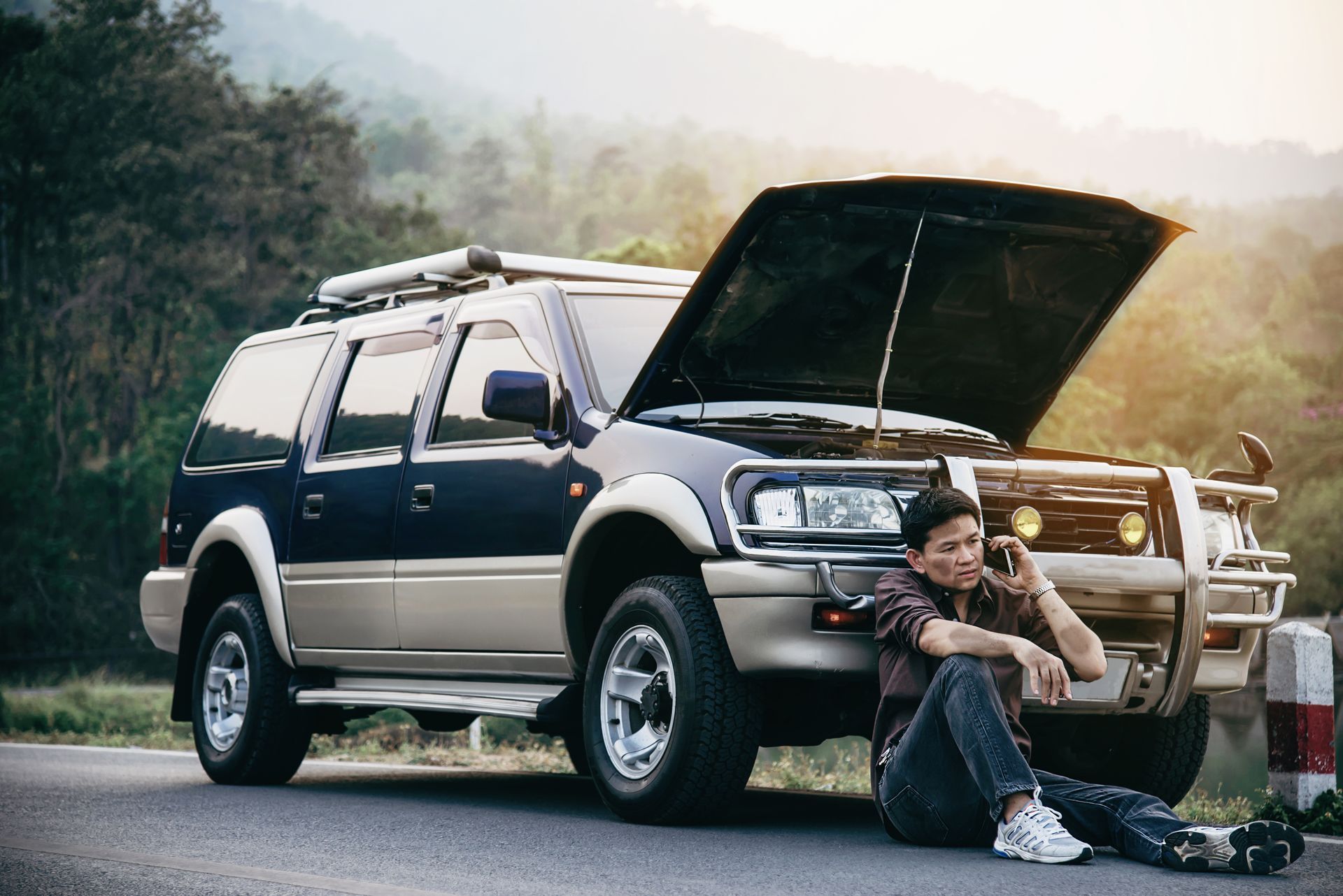SUV with hood open on roadside, man kneels beside it as smoke rises in a wooded mountain area