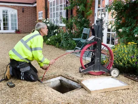 Worker in neon safety gear inspecting a drainage opening with a hose beside a red pump on a building courtyard.