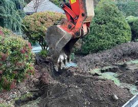 Excavator bucket digging soil beside shrubs and a driveway in a garden
