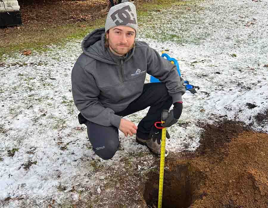 Person kneeling outdoors by a hole in snowy ground, holding a yellow measuring tape.
