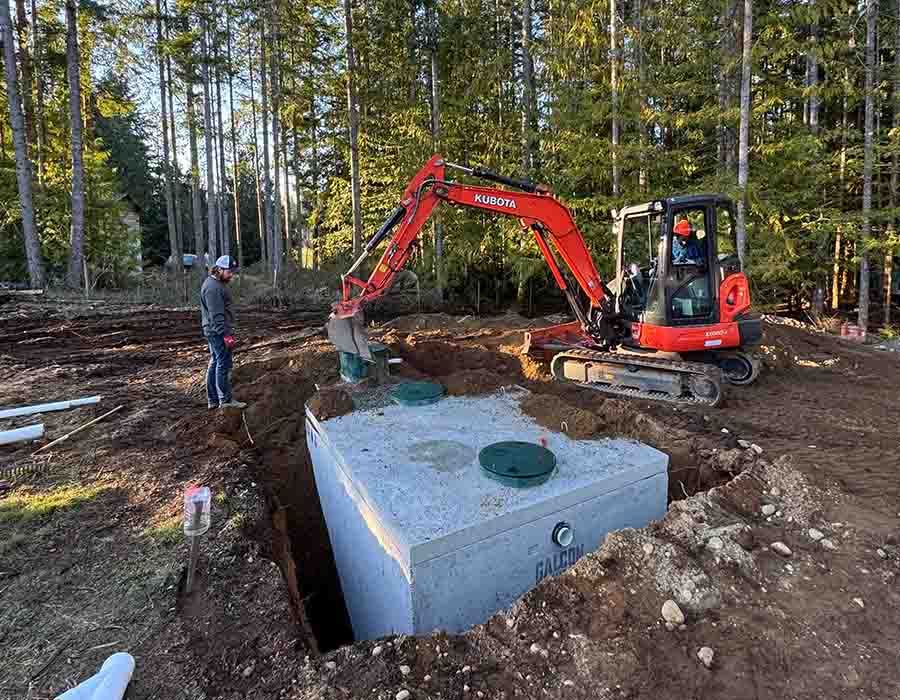 Excavator installing a concrete tank in a wooded construction site while a worker watches.