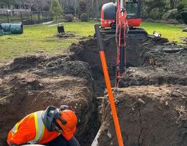 Construction worker in orange safety gear digging a trench beside an excavator