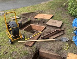 Open utility hole with tools and boards beside it on a muddy lawn
