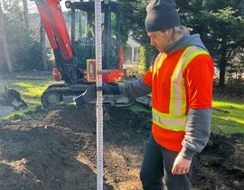 Worker in orange safety vest measuring ground beside a red excavator at a construction site
