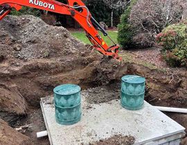 Orange Kubota excavator digging behind a concrete pad with two green vents in a landscaped yard