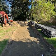 Backyard construction site with excavator, dirt path, and stacked stone blocks along a fence