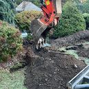 Excavator bucket digging up dark soil in a landscaped garden with shrubs and grass
