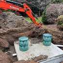 Orange Kubota excavator digging beside a concrete slab with two green barrels outdoors