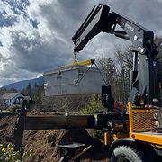 Crane lifting a gray metal container over a dump truck outdoors under cloudy skies