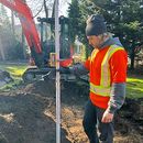 Worker in orange safety vest measuring ground beside a red excavator on a construction site