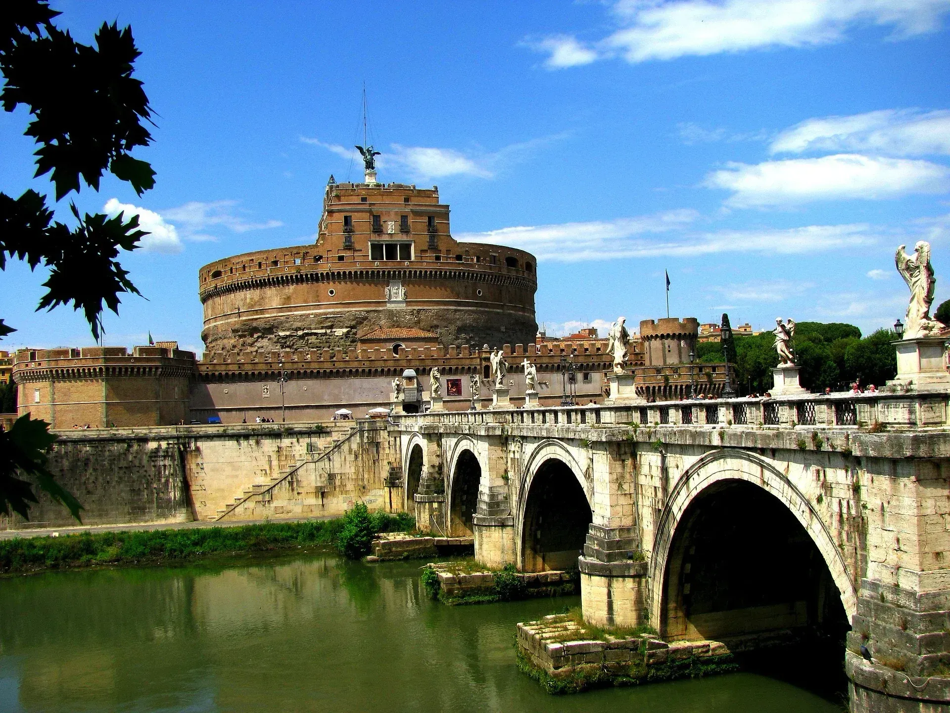 Stone bridge over a green river beside a historic round fortress under a blue sky