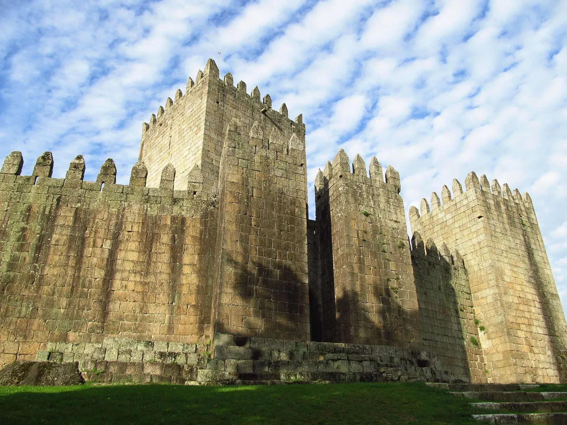Stone castle walls and towers under a blue sky, viewed from below on a grassy slope